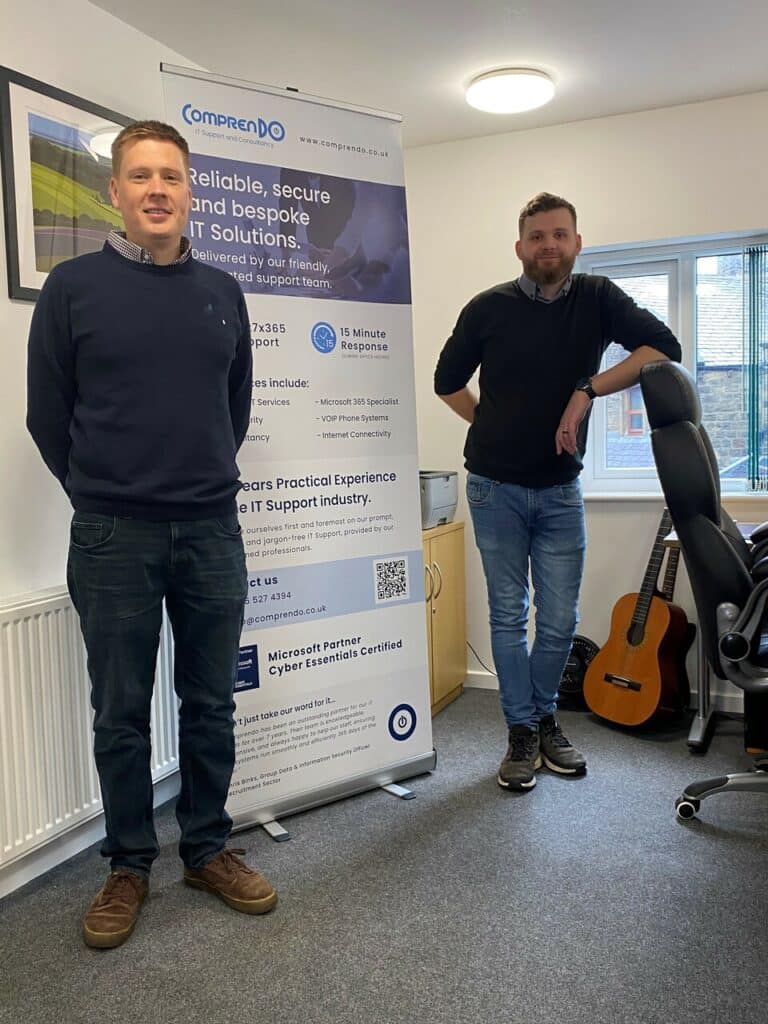 Daniel Hildred, Director of Comprendo, and Zack Thompson, IT technician, standing in the Comprendo office in front of a pop up banner.